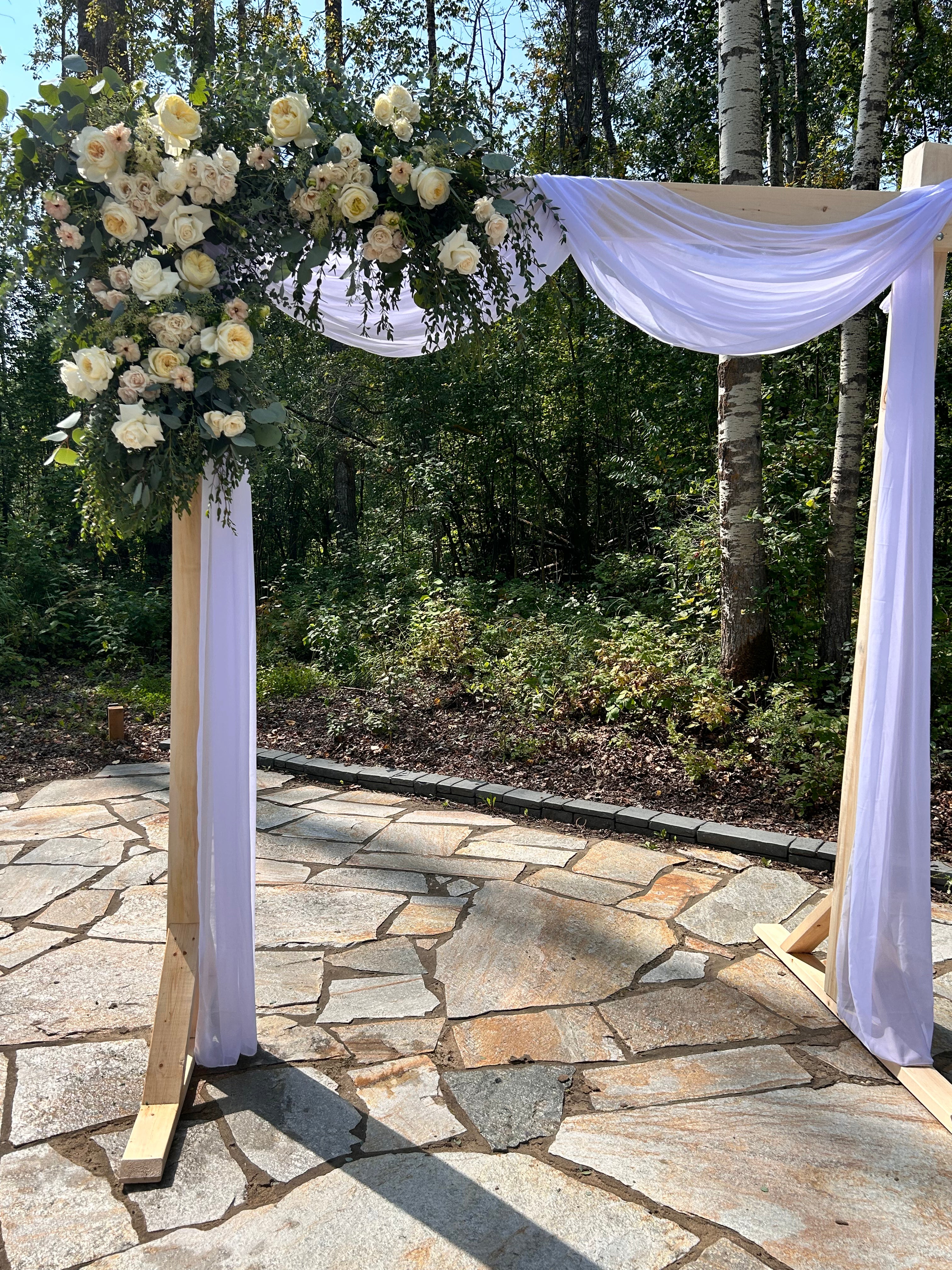 Decorative arch with white flowers and fabric on a stone pathway with trees in the background